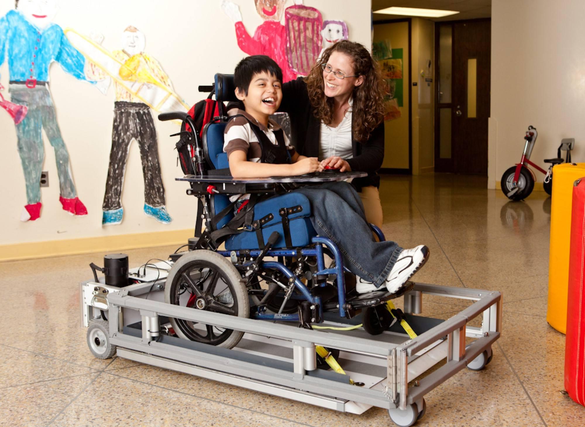 a woman kneels next to a child in a mobility aid